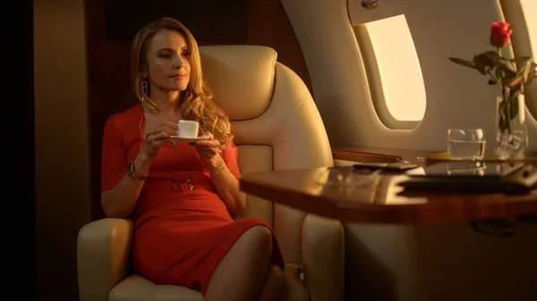 Passenger enjoy first class trip in golden sunlight. Rich woman in red dress drinking coffee on private airplane. Successful businesswoman relax check email social media. Wealthy blonde use smartphone