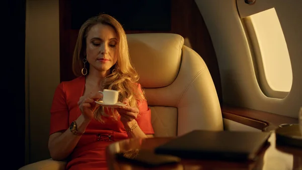 Tired entrepreneur resting airplane closeup. Beautiful blonde drinking coffee in golden sunlight. Pensive businesswoman lean chair recalling family happy moments. Wealthy woman looking jet window.
