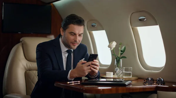 European businessman typing phone on airplane trip. Confident manager resting drinking coffee in luxury jet. Successful man texting surfing internet in suit. Happy gentleman check email use smartphone