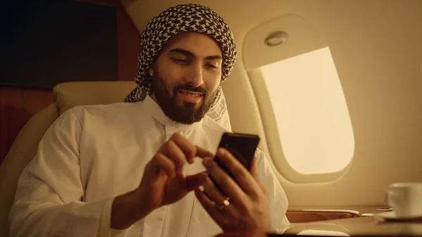 Rich man looking smartphone on travel closeup. Smiling arabian touching screen typing message on cellphone in luxury private jet. Cheerful bearded passenger browsing internet enjoying social media
