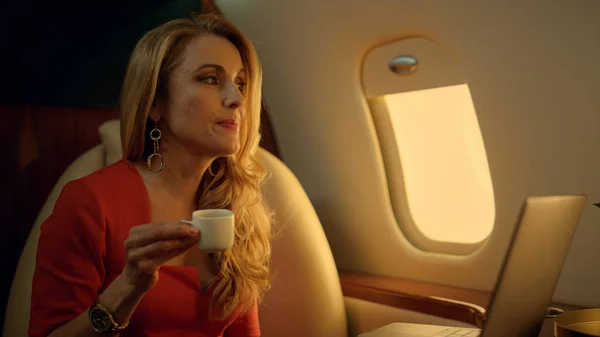 Smiling businesswoman traveling corporate jet closeup. Luxury lady resting alone chatting online. Relaxed elegant blonde use laptop computer drink coffee at airplane window. Wealthy lifestyle concept