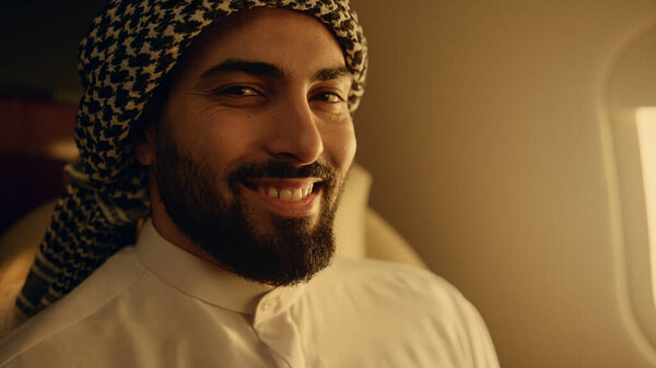 Улыбающийся арабик позирует у окна самолёта. Счастливый бизнесмен путешествует роскошь первого класса в традиционном наряде. Привлекательный бородатый владелец компании отдыхает наслаждаясь деловой поездкой. Богатые люди