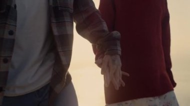 Unrecognizable woman and man walking on sea shore at sunrise. Closeup fashionable couple holding hands. Young hipsters wearing casual clothes. Sun lights shining between arms at sunset. Youth concept