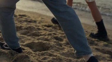 Closeup stylish couple feet walking on sandy beach. Unrecognizable girl and guy legs going along ocean shore. Young woman and man wearing fashionable sneakers and boots outdoors. Leisure concept