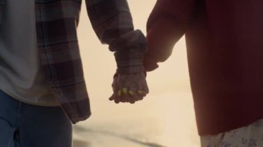 Unrecognizable couple holding hands at sea beach. Loving girl and guy walking along ocean shore at sunset. Closeup friends touching arms. Sun lights blinking between people hands. Friendship concept