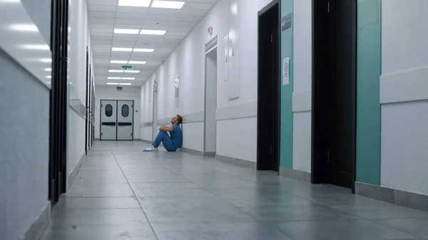 Unknown weary surgeon sitting floor modern clinic wearing blue uniform. Exhausted doctor staying on empty hospital corridor. Lonely tired man medicine worker resting leaning on wall hallway.
