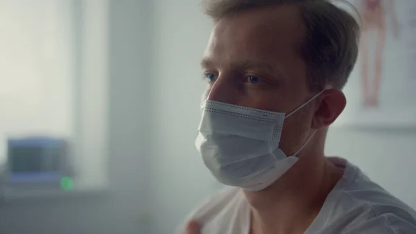 Tired patient sitting wearing mask in modern doctor office closeup. Young serious man holding arm after vaccine injection. Blond guy waiting physician in health clinic. Vaccination healthcare concept.