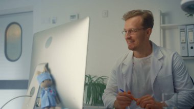 Gülümseyen adam doktorun klinikteki modern bilgisayarı kullanarak video konferansı var. Genç hekim covid salgını olan bir hastaya danışmanlık yapıyor. Arkadaş canlısı terapist hastanede online tanı koyuyor.