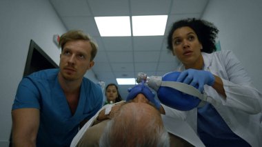 Endişeli tıbbi grup sedye hastane koridoruna hastayı itiyor. Stresli çok ırklı doktorlar hasta ihtiyarı acil servise taşıyorlar. Genç afro kadın sıhhiyeci ambu çantası kullanıyor..