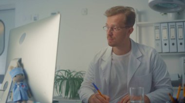 Ciddi bir doktor video konferansında reçete yazıyor. Modern kabineyi kapatıyorlar. Tıbbi konsültasyon için bilgisayarı kullanan profesyonel bir doktor. Erkek doktor uzaktan sağlık muayenesi yapıyor.