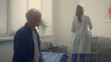 Klinikte tıbbi muayeneyi bekleyen maskeli yaşlı bir adam var. Bilinmeyen kadın doktor, kıdemli hasta için test koleksiyonu hazırlıyor. Emekliler kanepede, modern hastanede terapist ziyaretinde..