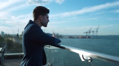 Thoughtful businessman looking sea on glass terrace. Handsome bearded guy lean thinking business decision in morning. Successful freelancer relaxing taking break on rooftop balcony at port cranes view