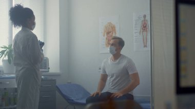 Hastalık belirtilerini tartışan çok ırklı doktor kadın hastane ofisinde oturuyor. Hemşire veri sonuçlarıyla birlikte tablet bilgisayarı tutuyor. Afrikalı Amerikalı doktor klinikte maskeli adama danışmanlık yapıyor..