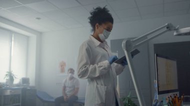 Maskeli kadın terapist tablet bilgisayardaki hasta bilgilerine bakıyor. Sağlık kliniğinde profesyonel tıbbi danışmanlık. Modern hastane ofisindeki Afrikalı Amerikalı doktor erkek sağlığı geçmişini kontrol ediyor..