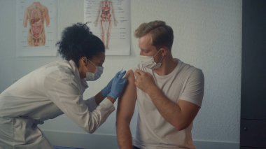 Modern klinikte koronavirüs aşısı olan bir adam. Afrikalı Amerikalı kadın doktor koruyucu maske takan genç müşteriyi aşılıyor. Sağlık hemşiresi omzuna iğne yapıyor..