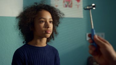 Güzel genç kız modern klinikteki nörologu ziyaret ediyor. Bilinmeyen doktor içeride nörolojik çekiç kullanarak göz refleksini test ediyor. Afrikalı Amerikalı genç pediyatrist kontrolünde kanepede oturuyor.. 