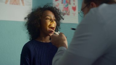 Cute teen girl opening mouth showing doctor throat in modern clinic close up. Pediatrician examining teenager using tongue depressor with flashlight. African american patient visit physician office.