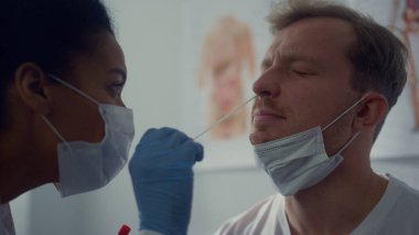 Profesyonel doktor burun hastasından örnek alıyor. Genç sarışın adam modern hastane ofisinde hızlı covid19 testinden geçiyor. Afrikalı Amerikalı doktor pamuk çubuğu kullanarak malzeme topluyor..