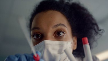 Kadın doktor, Coronavirus testi için pamuk çubuğu kullanarak örnek alıyor. Yüz maskesi takan Afrikalı neşeli hemşire klinikten hasta tükürüğü topluyor. Genç doktor tüpü tutuyor..