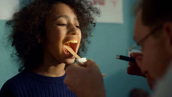 European doctor using tongue depressor checking girl throat close up. Man pediatrician looking in open mouth african american preteen with flashlight searching covid symptoms in hospital office.