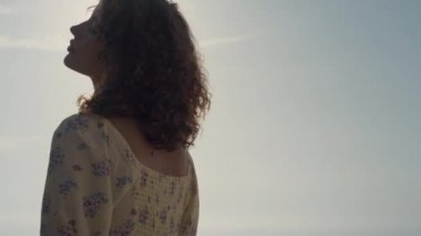Güzel genç bayan deniz sahilinde tek başına güneşin tadını çıkartıyor. Deniz kenarında poz veren kıvırcık, çekici bir kadın. Hoş bir elbise giymiş. Tasasız güzel kız doğada dinleniyor. Özgürlük kavramı.