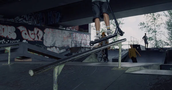 Millennial man jump on rail with scooter at city skate park. Close up extreme sportsmen riding on urban skatepark. Male friends performing tricks with skateboard and bmx bike outdoors. Action concept