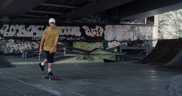 Young skater teenager riding on skate board at skate park with graffiti wall. Sporty male hipster in casual clothing skateboarding outdoors. Hobby active lifestyle youth culture concept.