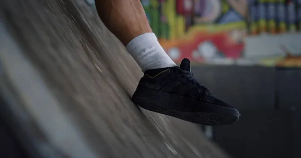 Unknown man having break after riding on skate board at skatepark. Closeup young skater relaxing at graffiti background. Close up unknown male legs in sneakers. Extreme sport recreation concept.