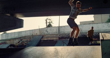 Teenage athletes performing stunt together at city skate park. Active people training jump trick at urban bright sunshine space. Extreme sport favorite hobby leisure modern young people concept. 