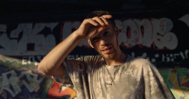 Kaykay parkındaki eğitimden sonra duvar yazısı geçmişi olan yakışıklı bir adam dinleniyor. Şehir kaykay parkında kameraya bakan yorgun adamı kapat. Aktif şehir alt kültür aktivitesi yaz konsepti.