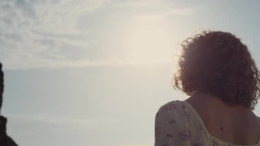 Bilinmeyen kıvırcık kadın, yüzünü güneş ışığına çeviriyor. Arka manzara genç, rahat kız dışarıda tek başına parlak sabah güneşinin tadını çıkarıyor. Doğada rahatlayan sevimli elbiseler giyen tanınmayan bir kadın..