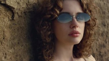 Pretty face young model looking camera confidently in sunglasses. Portrait of attractive sexy woman leaning on sand hill outdoors. Curly serious girl posing standing on seacoast alone. Relax on nature