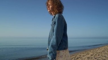 Attractive happy girl enjoying sunny day on sandy seacoast. Cute curly woman smiling camera having fun on ocean shore. Stylish positive lady wearing casual clothes posing on beautiful nature.
