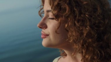 Gülümseyen, sakin, güzel bir kadının portresi. Şirin kıvırcık kız, kapalı gözlerle gün ışığına çıkıyor. Çekici genç bayan deniz sahilinde yalnız başına doğanın tadını çıkarıyor. Mutluluk kavramı.