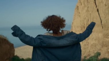 Deniz kıyısında boş kumsala giden genç bir kadın. Bilinmeyen aktif kız deniz kıyısında tek başına yürürken kollarını açıyor. Kıvırcık saçlı kadın kot ceket giyerek doğada rahatlıyor..