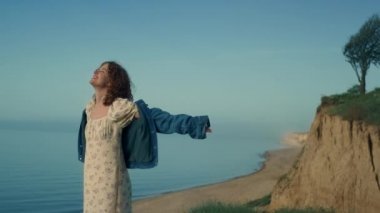 Happy calm woman enjoy nature on sand sea beach. Curly attractive girl spreading hands to sides standing empty seashore. Cute cheerful lady smiling raising face to sky outdoors. Nature beauty concept.