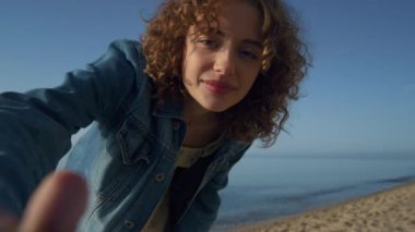 Deniz sahilinde kamerada gülümseyen mutlu güzel kadın. Yaklaş. Genç kıvırcık kız güneşli deniz kıyısında seyahat vlog 'u çekerken el sallıyor. Tarz sahibi pozitif bayan okyanus kıyısında rahatlar Güzel bir bahar günü