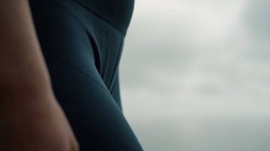 Bilinmeyen zayıf bir kadın koyu renk spor giyim içinde ayakta duruyor. Gergin göbek bacakları, bulutlu gri gökyüzünde tanınmayan bir kız. Deniz kıyısında tayt giyen atletik bayan eğitimi. Spor konsepti 