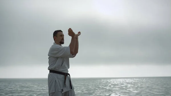 跆拳道人在平静的大海附近训练体力 留着胡子的运动员穿着阴天和服练习武术 以强壮的战士为重点 在户外进行双手操练 — 图库照片
