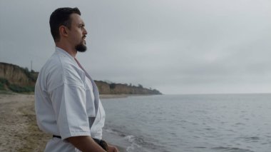 Kumsalda kimono giyerek karate egzersizi yapan sporcu. Yakından. Deniz kenarındaki bulutlu bir sabahta güçlü bir avcı uçağı eğitimi. Dışarıda tekvando antrenmanı yapan sakallı sporcu profili..