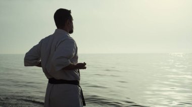 Arka plan atletik adam karate tekniğini biliyor sahilde günbatımının tadını çıkarıyor. Sakin deniz akşamları dövüş sanatları yapan güçlü bir dövüşçü. Tanınmayan sporcu kimono antrenmanı yapıyor..
