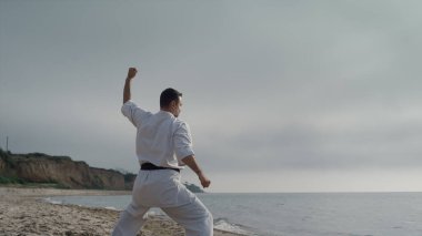 Kumlu kumsal güneşli sabahında karate dövüşçüleri antrenman yapıyor. Atletik adam dövüş pozisyonunda dışarıda dövüş sanatları çalışıyor. Genç sporcu, doğa üzerinde savaş egzersizleri yapıyor..