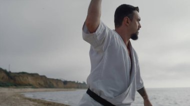 Kumlu plaj bulutlu sabahında dövüş sanatları pratiği yapan sakallı kung fu 'cu. Sportif sporcu dövüş egzersizleri açık havada kapalı hava. Kimono giyen genç bir karate dövüşçüsü..