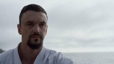 Güçlü sakallı adam deniz kıyısında kimono giyerek karate yumrukları yapıyor. Kumlu plaj bulutlu sabahında aktif sporcu eğitimi. Odaklanmış dövüşçü dışarıda hızlı hareket ediyor.