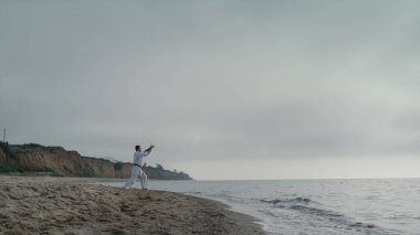 Karate sporcusu kumlu plaj bulutlu sabahında atak pratiği yapıyor. Deniz kıyısında dövüş sanatları eğitimi alan güçlü bir adam bulutlu bir havada. Aktif sporcu doğa üzerinde savaş egzersizleri yapıyor. Refah kavramı.