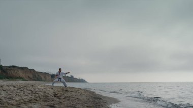 Aktif adam deniz kıyısında hava bulutlu bir günde dövüş tekniği eğitimi veriyor. Kumsalda kimono giyerek karate antrenmanı yapan sporcu. Güçlü sporcu dışarıda dinamik egzersizler yapıyor. Sağlıklı yaşam tarzı.