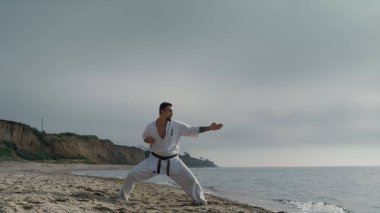 Karate sporcusu güneşli sahilde kimono giyerek tekme antrenmanı yapıyor. Güzel bir yaz sabahı sporcu dövüş sanatları eğitimi alıyor. Aktif sporcu dövüş yeteneklerini açık havada geliştirir. Spor konsepti.