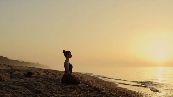 Focused woman practicing meditation on sandy beach summer evening. Yoga woman sitting lotus position near ocean waves. Attractive sportswoman making relaxing asana outdoors. Self care concept.