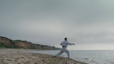 Aktif sporcu, güzel plaj manzaralı bulutlu bir günde karate egzersizleri yapıyor. Güçlü sakallı adam doğada dövüş tekniğini geliştirir. Kumlu tepelerde dövüş sanatları eğitimi alan genç bir sporcu..