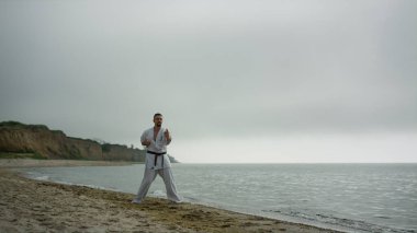 Judo dövüşçüsü kumlu sahilde beyaz kimono giyerek antrenman yapıyor. İnsanoğlu, sakin okyanus yakınlarında savaş tekniklerini öğreniyor. Güçlü sakallı sporcu deniz kenarında tekvando çalışıyor..